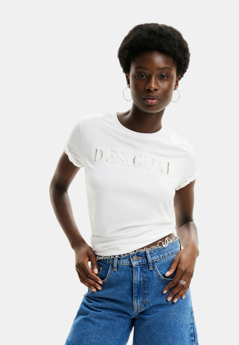 Camiseta blanca con letras en relieve "DESIGUAL", de manga corta, combinada con jeans de denim azul de cintura alta y accesorios plateados.