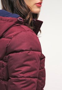 Bordowa kurtka puchowa o teksturowanym wykończeniu, z kapturem wyściełanym granatem i zatrzaskami, prezentująca pikowane detale ocieplające.