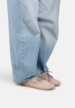 Vaqueros azul claro de pierna ancha combinados con zapatos estilo mocasín beige con cordones estampados y suelas marrón claro sobre un fondo blanco.