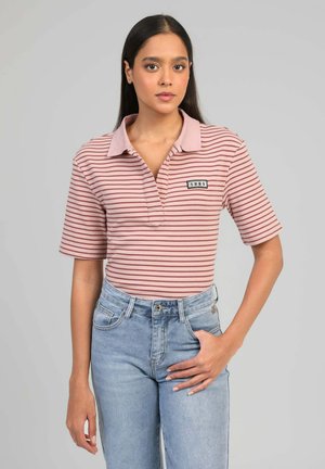 Jeune femme portant un polo rayé rose et blanc avec un patch "1985", un jean taille haute bleu clair, debout devant un fond gris uni.