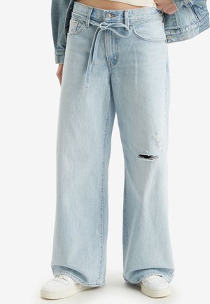 Lichtblauwe wijde jeans met een stoffen riem en een gescheurd detail aan de rechterdij, gedragen met witte sneakers en een spijkerjack.