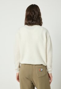 Ženska z rjavimi lasmi do ramen, oblečena v belo puhasto jopico in olivno zelene hlače z majhno pisano etiketo na zadnjem žepu.