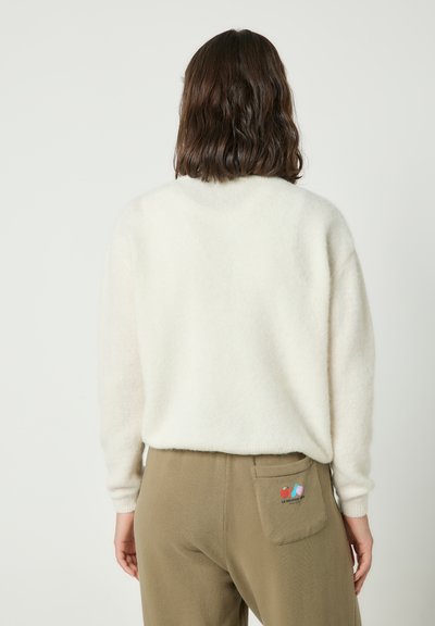 Femme aux cheveux bruns mi-longs portant un pull blanc en tissu duveteux et un pantalon vert olive avec un petit patch coloré sur la poche arrière.