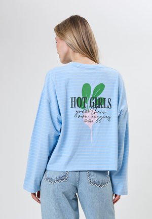 Kvinde iført en lyseblå stribet langærmet skjorte med teksten "Hot Girls grow their own veggies" og en radise grafik på ryggen.