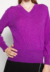 Pull violet à col en V avec manches longues, texture et tissu côtelés, présentant un ourlet ajusté et une finition lisse.