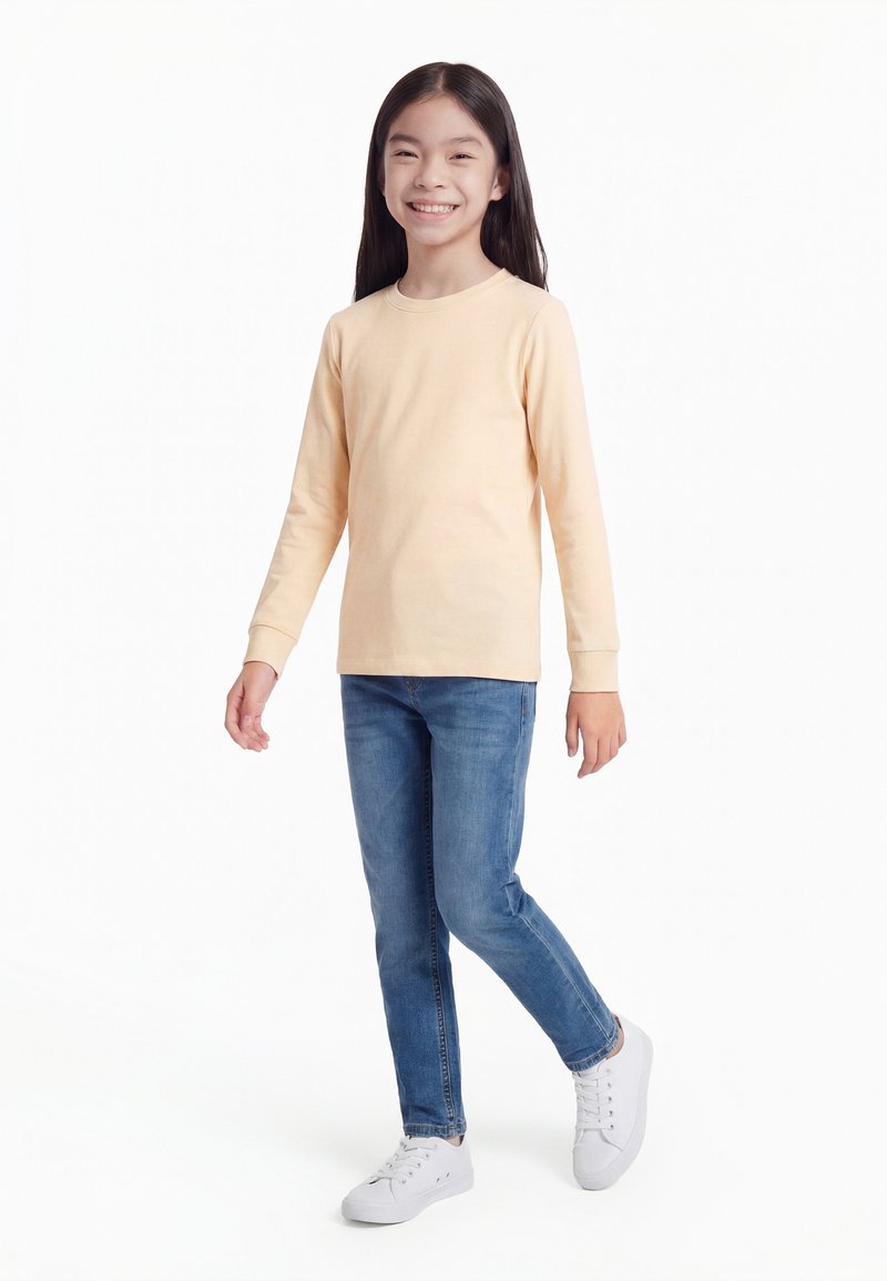 Niña sonriente con cabello largo oscuro, de pie, vestida con una camiseta beige de manga larga, jeans azules y zapatillas blancas contra un fondo blanco.