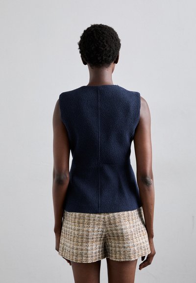 Mörkblå ärmlös topp med texturerad tyg och figurnära design, kombinerad med korta, rutiga shorts i brunt och kräm, sedd bakifrån.