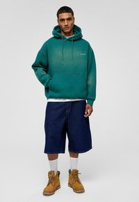 Groene gradient hoodie met een voorzak, gecombineerd met donkere denim shorts en gele werkschoenen. Witte sokken zichtbaar boven de schoenen.