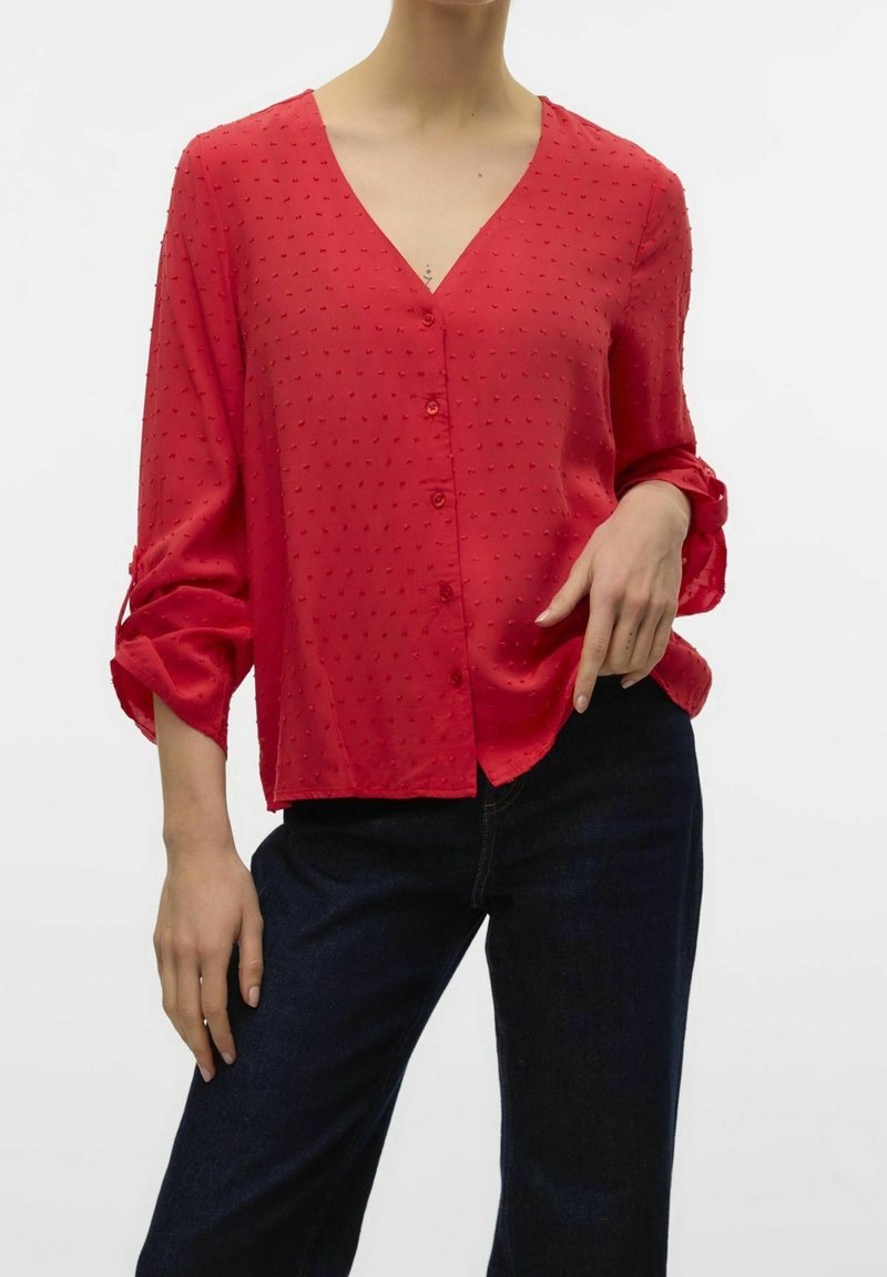 Femme portant un chemisier rouge texturé à boutons avec manches retroussées et un jean bleu foncé sur fond blanc.