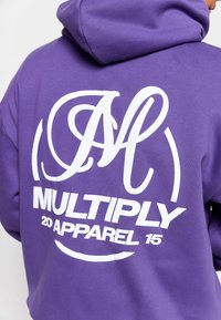 Lila Kapuzenpullover aus Baumwolle mit einem großen weißen Grafik auf dem Rücken, das stilisierte Buchstaben "M" sowie den Text "MULTIPLY 20 APPAREL 15" zeigt.