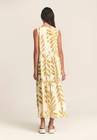 Dolga obleka brez rokavov v beli barvi z rumenimi listnatimi vzorci, slojevitim dizajnom in mehko teksturo. Nošena z lahkimi čevlji z blok peto.