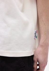 Ανοιχτό μπεζ βαμβακερό t-shirt, με πλαϊνή ετικέτα με πράσινες, λευκές και κόκκινες λεπτομέρειες, και ευθεία ρίγα με ορατή ραφή.