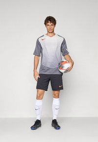 Γκρι και άσπρη αθλητική φανέλα ποδοσφαίρου με υφή σχεδίου, μαύρο σορτς, άσπρες κάλτσες και μαύρα ποδοσφαιρικά παπούτσια, κρατώντας μια ποδοσφαιρική μπάλα με σχέδιο.