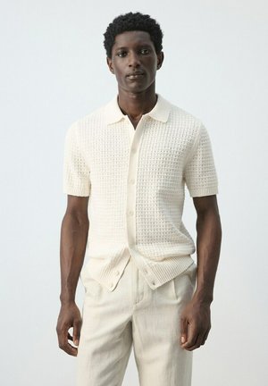 Homme portant une chemise en maille crème à manches courtes avec des boutons et un pantalon beige clair, debout devant un fond clair uni.