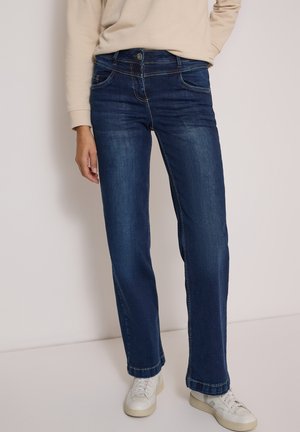 Person trägt dunkelblaue Jeans mit geradem Bein, beige langärmliges Oberteil und weiße Sneakers, steht vor einem schlichten hellen Hintergrund.