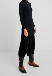 Robe noire à manches longues avec un col zippé, associée à un pantalon plissé noir et des bottes de cowboy marron avec des accents noirs et bruns.