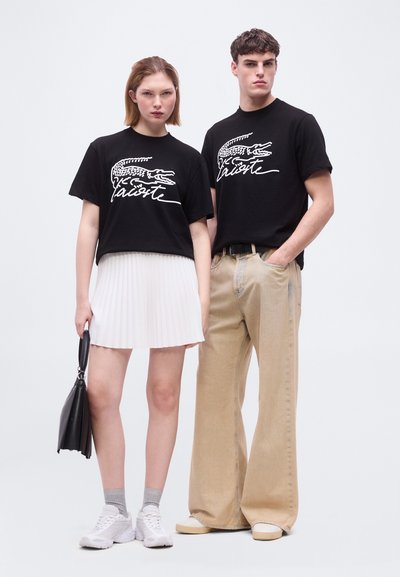 Млада жена и мъж стоят един до друг, облечени с черни тениски Lacoste, бяла пола и бежови панталони, с ежедневни обувки и неутрални изражения на лицата.