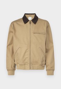 Chaqueta bomber color nude con cuello marrón oscuro, cierre frontal con cremallera, bolsillos laterales y un bolsillo en el pecho. Fabricada en un tejido resistente.