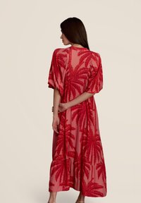 Robe maxi en tissu transparent corail avec un motif de feuilles de palmier rouge. Des manches bouffantes et un ourlet en cascade ajoutent du volume à la conception.