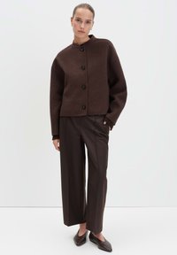 Vrouw in een donkerbruine, met knopen sluitende cardigan, bijpassende brede broek en puntige flats staand tegen een effen lichte achtergrond.