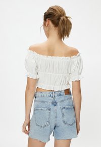 Biała, odsłonięta ramiona crop top z teksturowanej tkaniny i falbaniastymi brzegami, w zestawie z jasnoniebieskimi dżinsowymi szortami z postrzępionymi końcami i tylnymi kieszeniami.