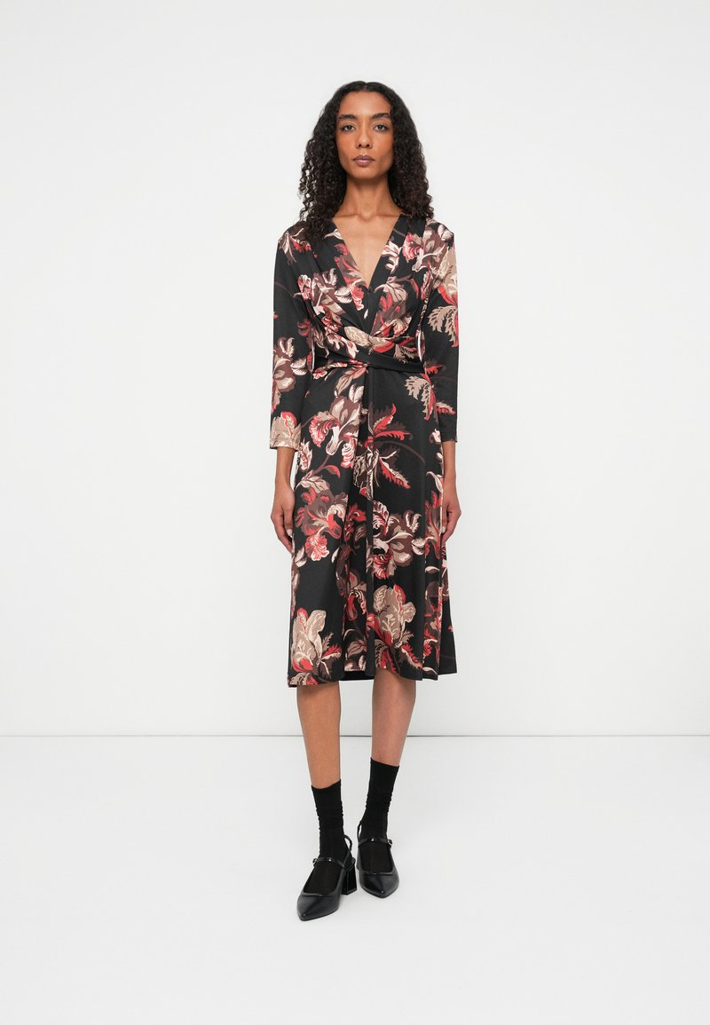 Vrouw met krullend haar draagt een zwart bloemen midi-jurkje met lange mouwen en zwarte sokken met zwarte hakken, staand tegen een witte achtergrond.