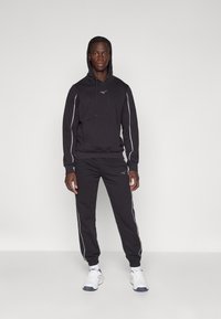 Black athletic hoodie with drawstrings and logo, paired with matching joggers featuring white side stripes, worn with white sneakers.