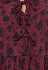 Abito color bordeaux in tessuto testurizzato, decorato con motivi floreali neri. Legatura al collo con una piccola apertura a forma di chiave e una vita arricciata.