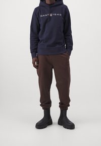 Persona con sudadera azul marino con el logo "GANT 1949", pantalones jogger marrones y botas negras gruesas, de pie contra un fondo claro y liso.
