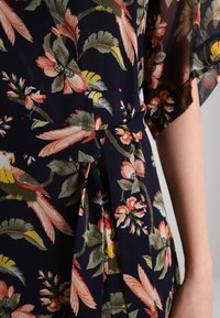 Närbild av marinblå blommig klädseltyg med rosa, gula och gröna blad och blommor, med ett knytbälte i midjan och genomskinliga ärmar.