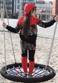 Czerwony i czarny dres z kwiatowym nadrukiem i kapturem. Stoi na okrągłej huśtawce, otoczony łańcuchami i piaszczystym placem zabaw.