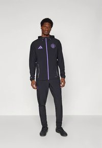 Zwarte sportieve trainingspak met een jas met ritssluiting, paarse accenten en een logo, gecombineerd met bijpassende broek. Lichtgewicht, sportief ontwerp.