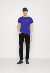 Blauwe katoenen t-shirt met ronde hals, zwarte denim jeans en zwarte sneakers. Model draagt een zwarte pet. Eenvoudig ontwerp zonder patronen.