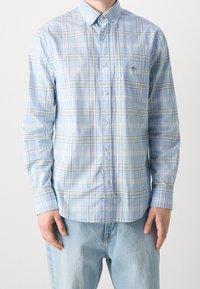 Camicia a quadri azzurri chiari con colletto abbottonato, maniche lunghe e taschino sul petto. Presenta linee bianche e viola che si intersecano.