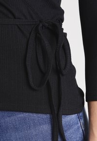 Černý žebrovaný top s designem do zavinování, se šňůrkami v pase. Vyrobeno z elastického materiálu; zobrazeno s modrými džíny.
