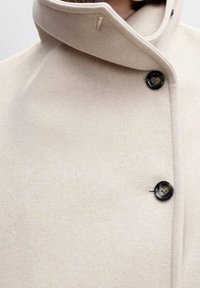 Cappotto di lana beige con colletto alto, caratterizzato da un unico bottone nero e una superficie liscia. Vista ravvicinata del colletto e dei bottoni.