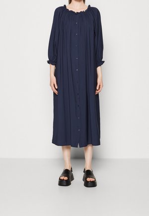 Robe midi bleu marine avec un col rond froncé, un devant boutonné, des manches longues avec poignets élastiques, et un tissu doux. Sandales à plateforme noires.