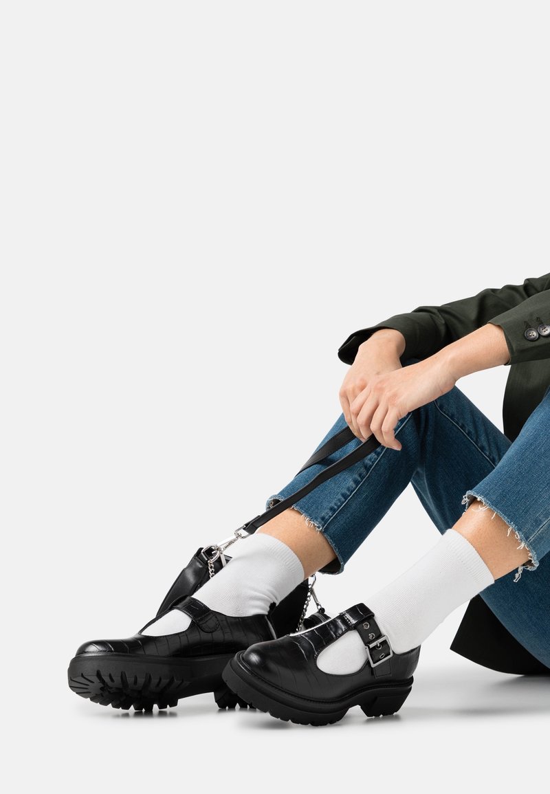 Chaussures en cuir noir avec une finition texturée, des accents de boucle et une semelle robuste. Portées avec des chaussettes blanches et un jean bleu effiloché.