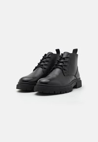 Botines de cuero negro con un acabado liso, puntera redonda y cordones en la parte delantera. Suela de goma negra gruesa con textura en la parte inferior. Lengüeta trasera.