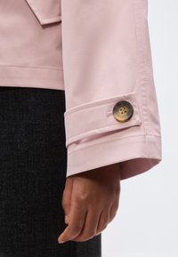 Temnopolta roka, ki počiva ob svetlo rožnati rokavici jakne z detajlom gumba, na temnem tkaninskem ozadju.