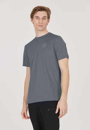 Jonge man met een grijs shirt met korte mouwen en een zwarte broek, staand tegen een eenvoudige lichte achtergrond.