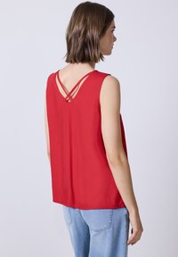 Top rojo sin mangas con espalda en V y tirantes cruzados; hecho de una tela suave. Combinado con jeans de tiro alto en azul claro.