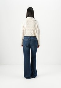 Pull beige en tricot avec un large col. Jean bleu évasé à taille mi-haute, avec des poches arrière et des détails de couture foncés.