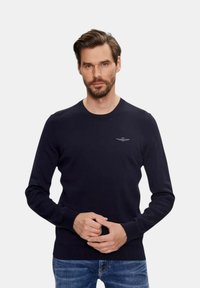 Navyblauwe gebreide trui met een ronde hals, geribbelde manchetten en zoom, met een subtiele logo op de borst. Gemaakt in combinatie met denim jeans.