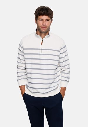 Hombre con un pulóver blanco de media cremallera con rayas horizontales grises y pantalones oscuros, de pie con las manos en los bolsillos contra un fondo liso.