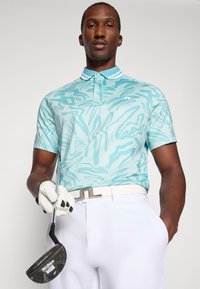 Polo azul-claro com padrão abstrato, mangas curtas e colarinho branco, combinado com calças brancas e segurando um taco de golfe.