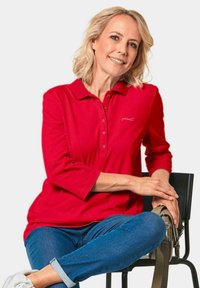 Rode polo met lange mouwen, voorzien van een kraag en knoopsluiting. Heeft een subtiele logo op de linkerborst, gecombineerd met blauwe jeans en een schoudertas.