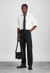 Cămașă albă cu mânecă scurtă, cravată neagră, pantaloni negri pliați și pantofi din piele negri. Ținând o geantă de mână neagră. Ținută simplă, formală.