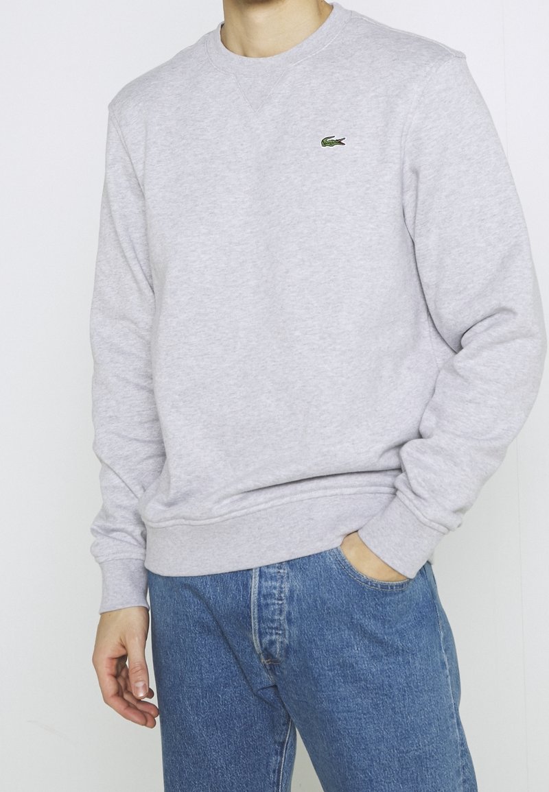 Felpa grigia chiara con un tessuto texture, polsini e orlo a costine, con un piccolo logo ricamato verde sul petto. Abbinata a jeans blu.