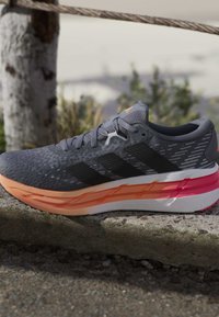 Chaussure de course grise et noire avec semelle orange et rose posée sur une surface en pierre rugueuse à l'extérieur près d'une clôture en corde et d'un tronc d'arbre.